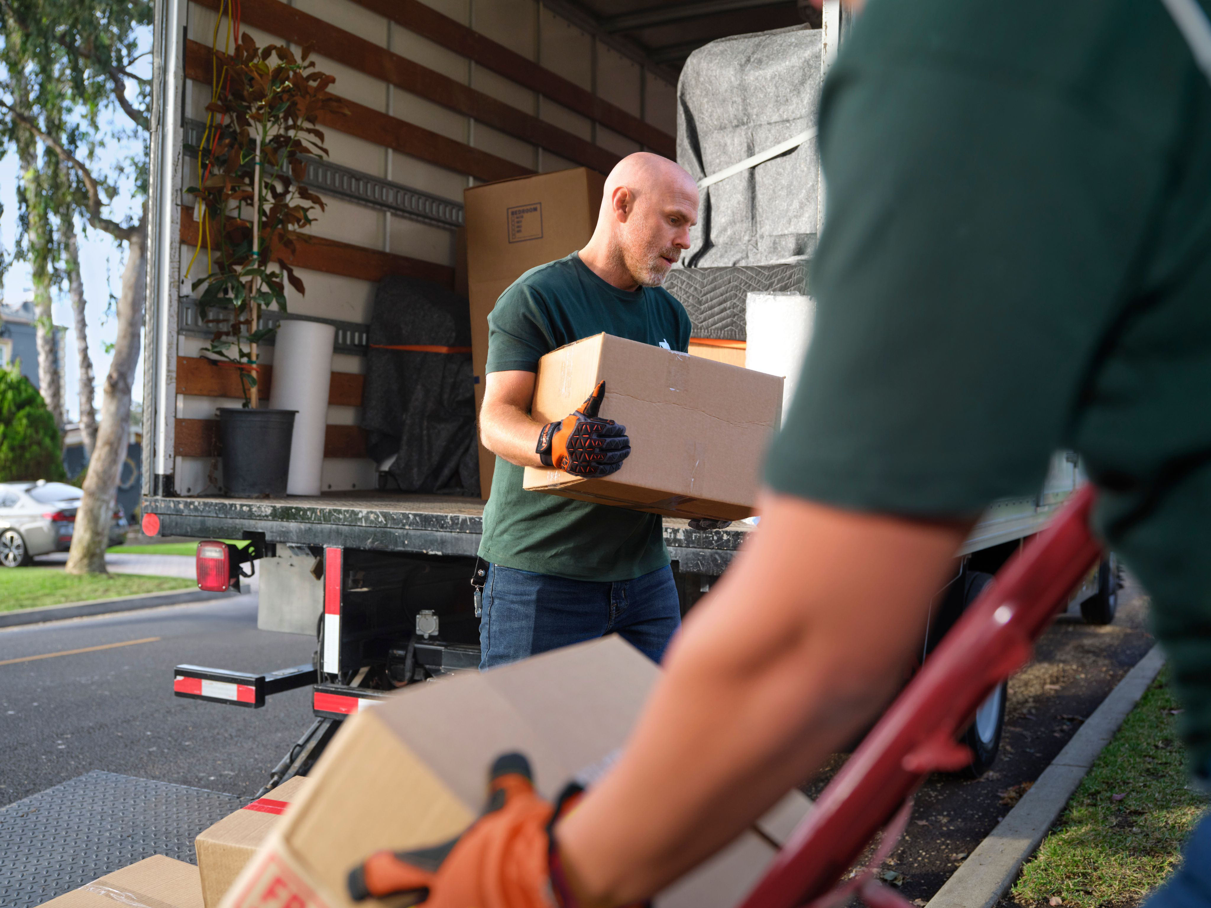 Two movers loading cardboard boxes onto a truck, with one man lifting a box inside while the other uses a hand truck in the foreground.